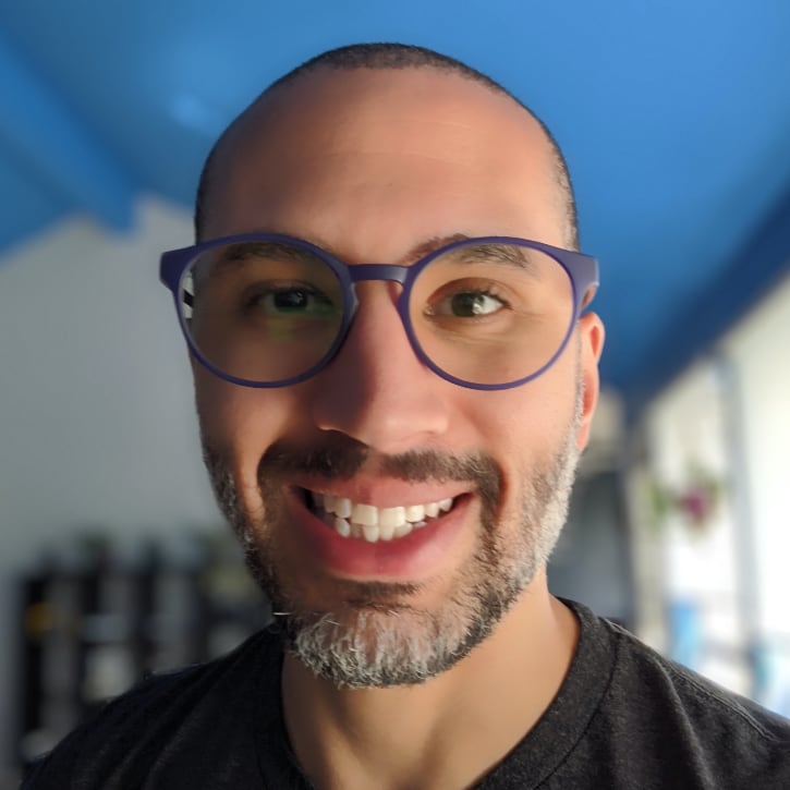 Photograph of Rob Lowe's head and shoulders. He is wearing a dark grey shirt, blue glasses, and a beard. The background is blurry, however a white wall and blue ceiling are recognizable.